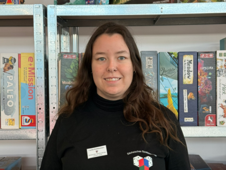 Carina Klinkhammer im Sächsischen Spielezentrum, Ludothek Leipzig, mit schwarzem T‑Shirt vor einem Brettspiele-Regal.