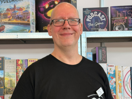 Carina Klinkhammer vom Sächsischen Spielezentrum, Ludothek Leipzig: Marcus mit schwarzem T-Shirt und Brille vor Spielregalen.