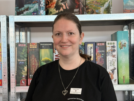 Carina Klinkhammer vom Sächsischen Spielezentrum trägt ein schwarzes T‑Shirt und steht vor einem Regal mit Brettspielen in der Ludothek Leipzig.