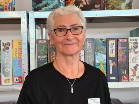 Carina Klinkhammer mit Brille und grauem Kurzhaar vor Brettspielregalen der Ludothek Leipzig im Sächsischen Spielezentrum, mit schwarzem Shirt.