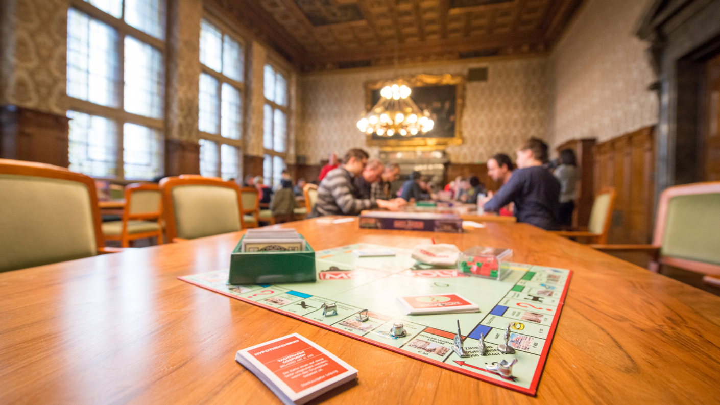 Ein Monopoly-Spielbrett mit Spielfiguren und Karten auf einem Holztisch in einem Raum mit vielen Personen im Hintergrund.