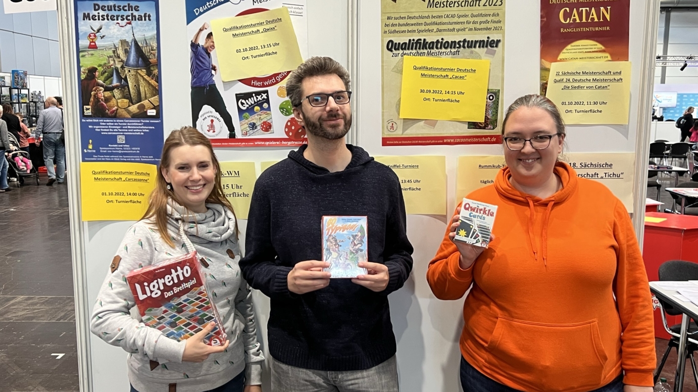 Drei Personen halten Brettspiele in der Hand und stehen vor einer Wand mit verschiedenen Plakaten auf einer Messe.