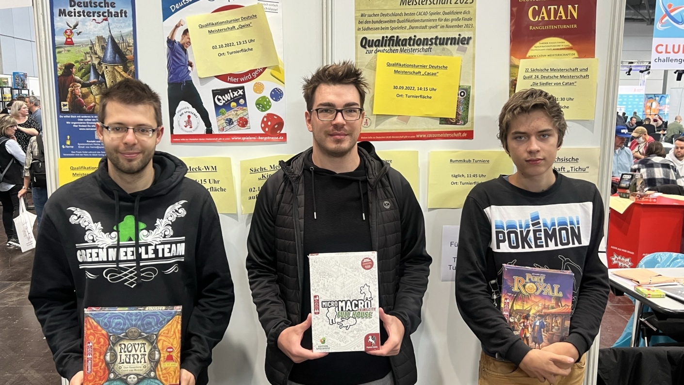 Drei Personen halten Brettspiele in ihren Händen, während sie vor einem Messestand stehen.