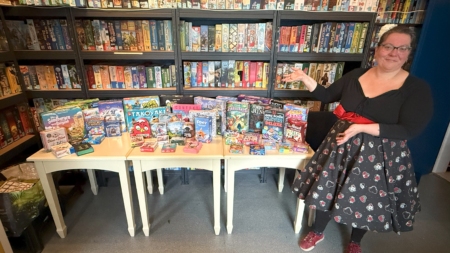 Frau zeigt eine Auswahl an Brettspielen auf einem Tisch vor einem Bücherregal.