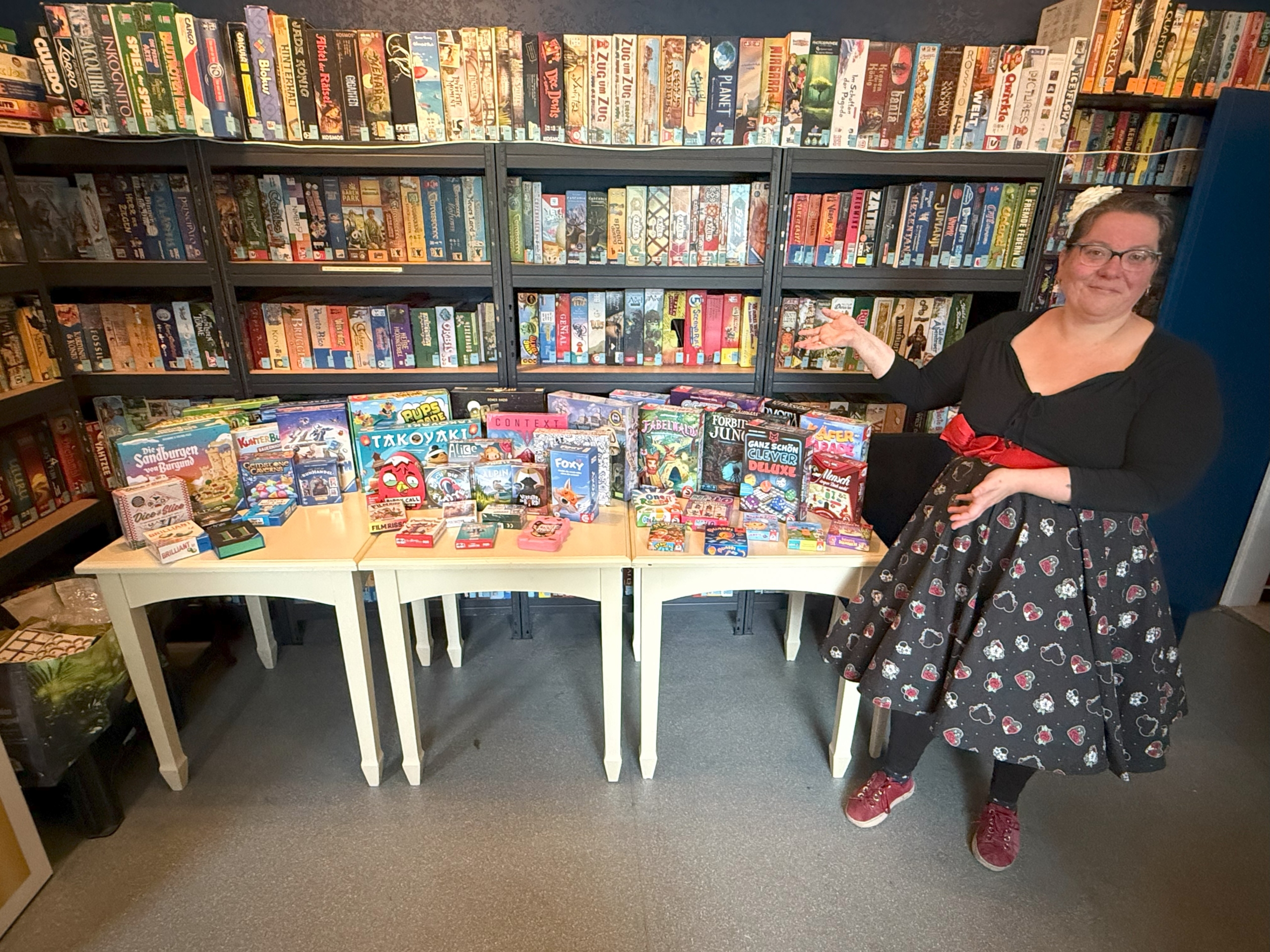 Frau zeigt eine Auswahl an Brettspielen auf einem Tisch vor einem Bücherregal.