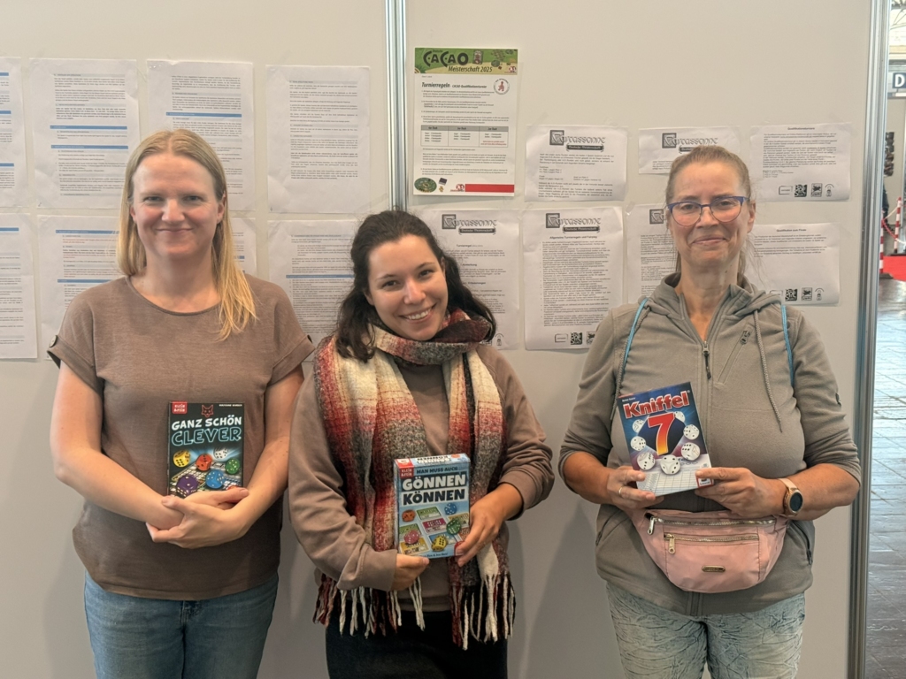 Drei Frauen halten verschiedene Gesellschaftsspiele in der Hand, stehen vor einer Wand mit Informationsblättern.