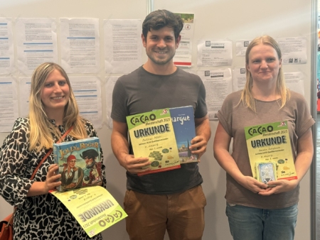 Drei Personen halten Urkunden und Spiele in den Händen auf einer Messe.