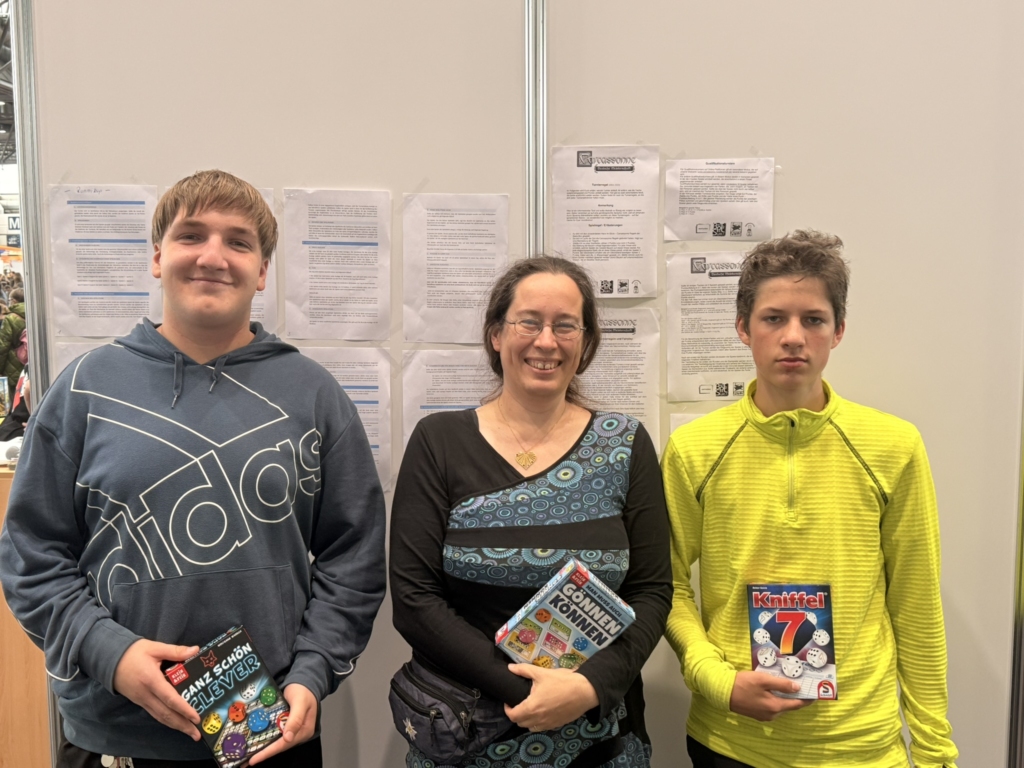 Drei Personen stehen nebeneinander und halten Bücher in der Hand, vor einer Wand mit Informationsblättern.