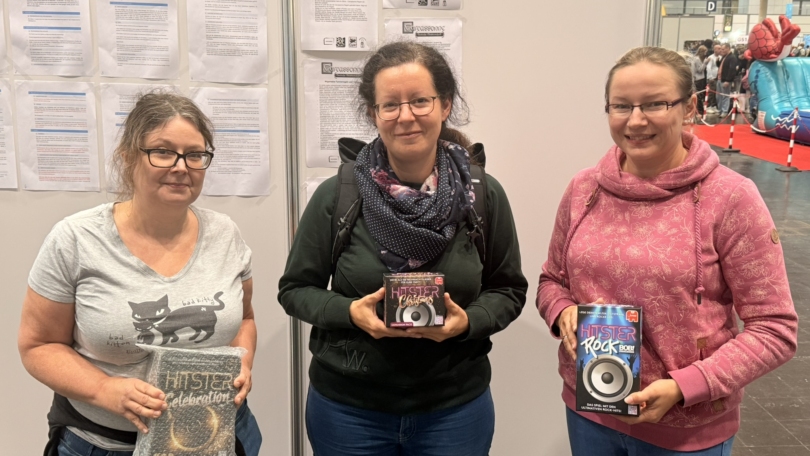 Drei Frauen halten Brettspiele in der Hand, während sie vor einer Wand mit Informationsblättern stehen.