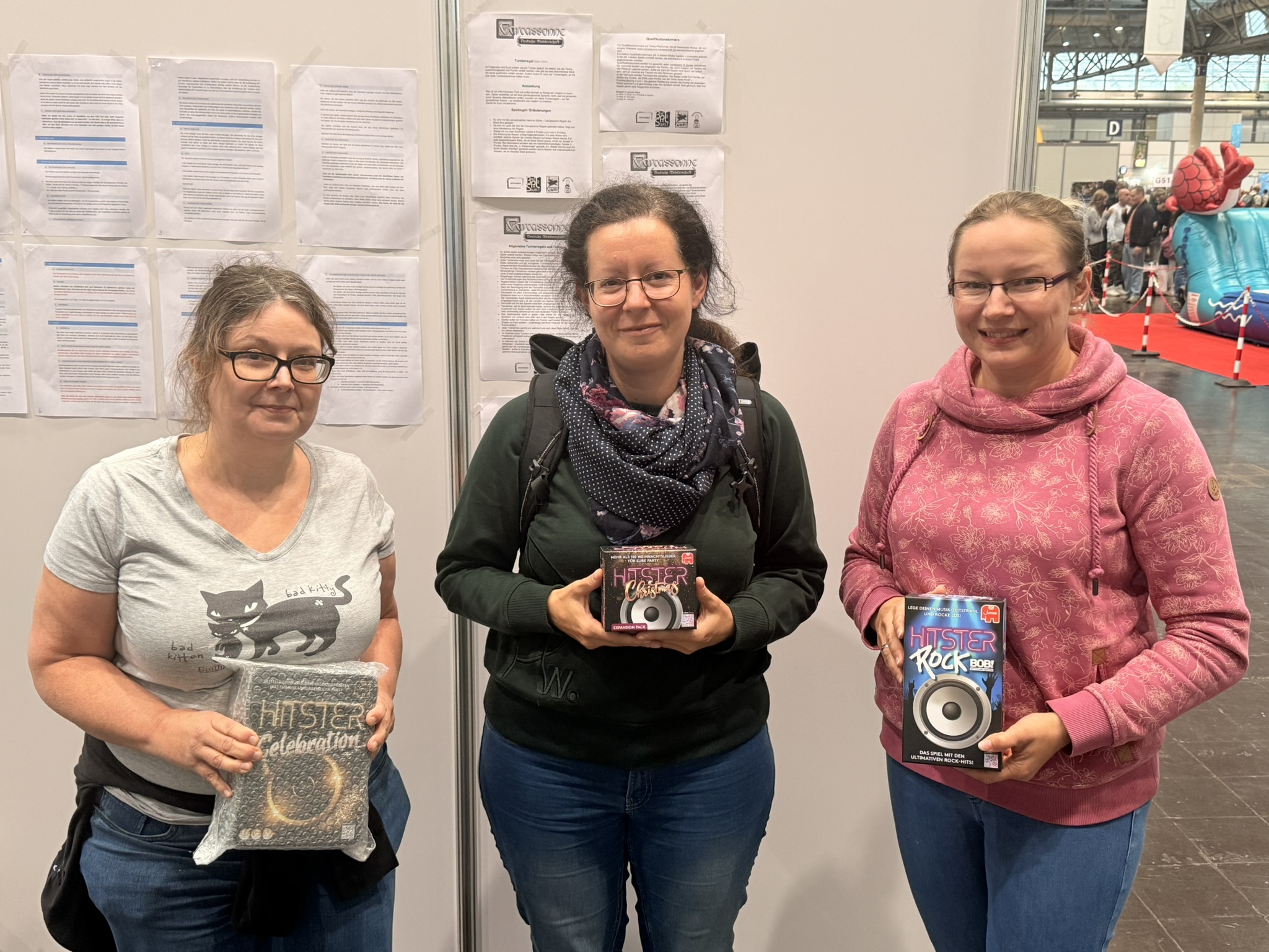 Drei Frauen halten Brettspiele in der Hand, während sie vor einer Wand mit Informationsblättern stehen.