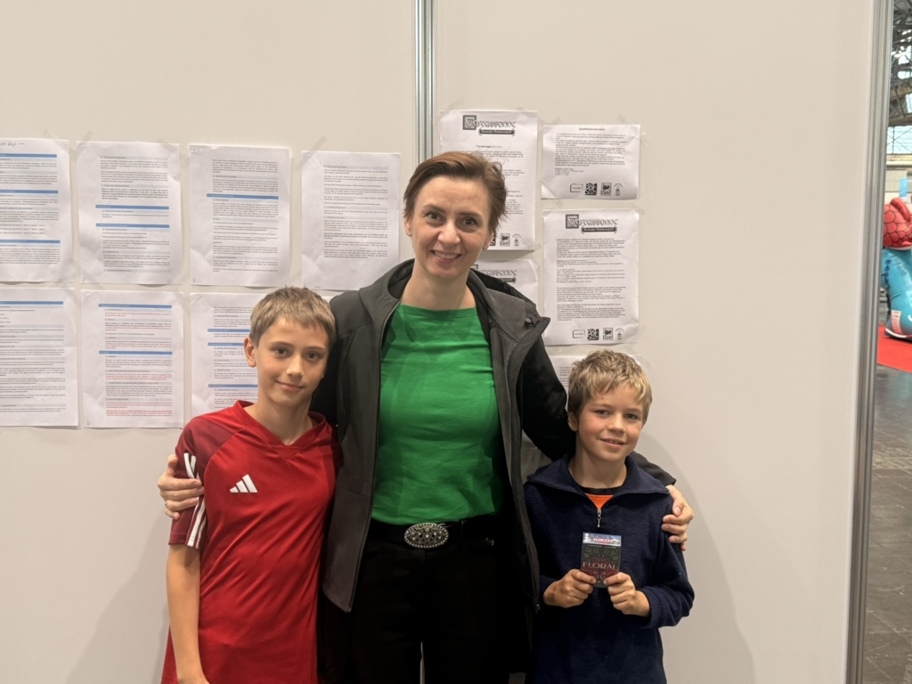 Eine Frau steht zwischen zwei Jungen, die an einer Wand mit Informationsblättern posieren.