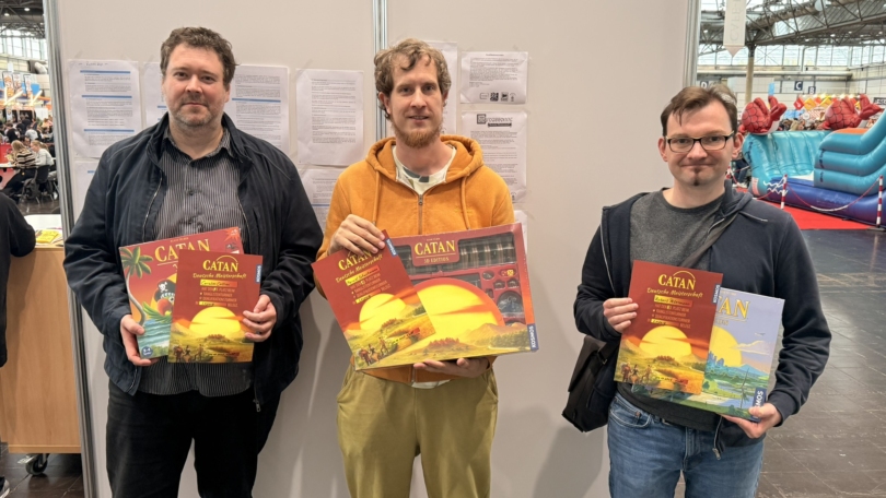 Drei Männer halten verschiedene Editionen des Brettspiels Catan in den Händen.