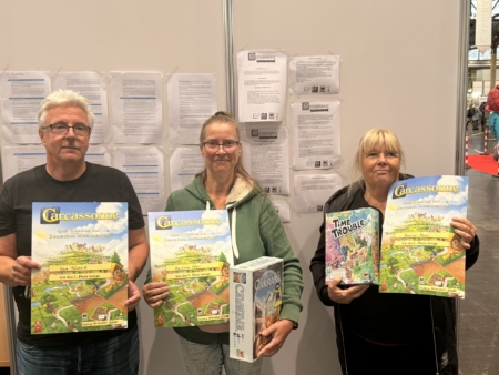 Drei Personen halten Brettspiele, darunter Carcassonne und Time Trouble, auf einer Messe.