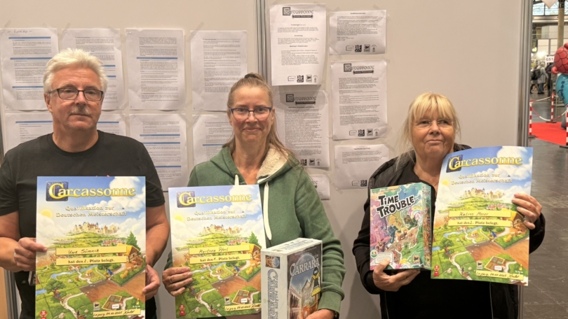 Drei Personen halten Brettspiele, darunter Carcassonne und Time Trouble, auf einer Messe.