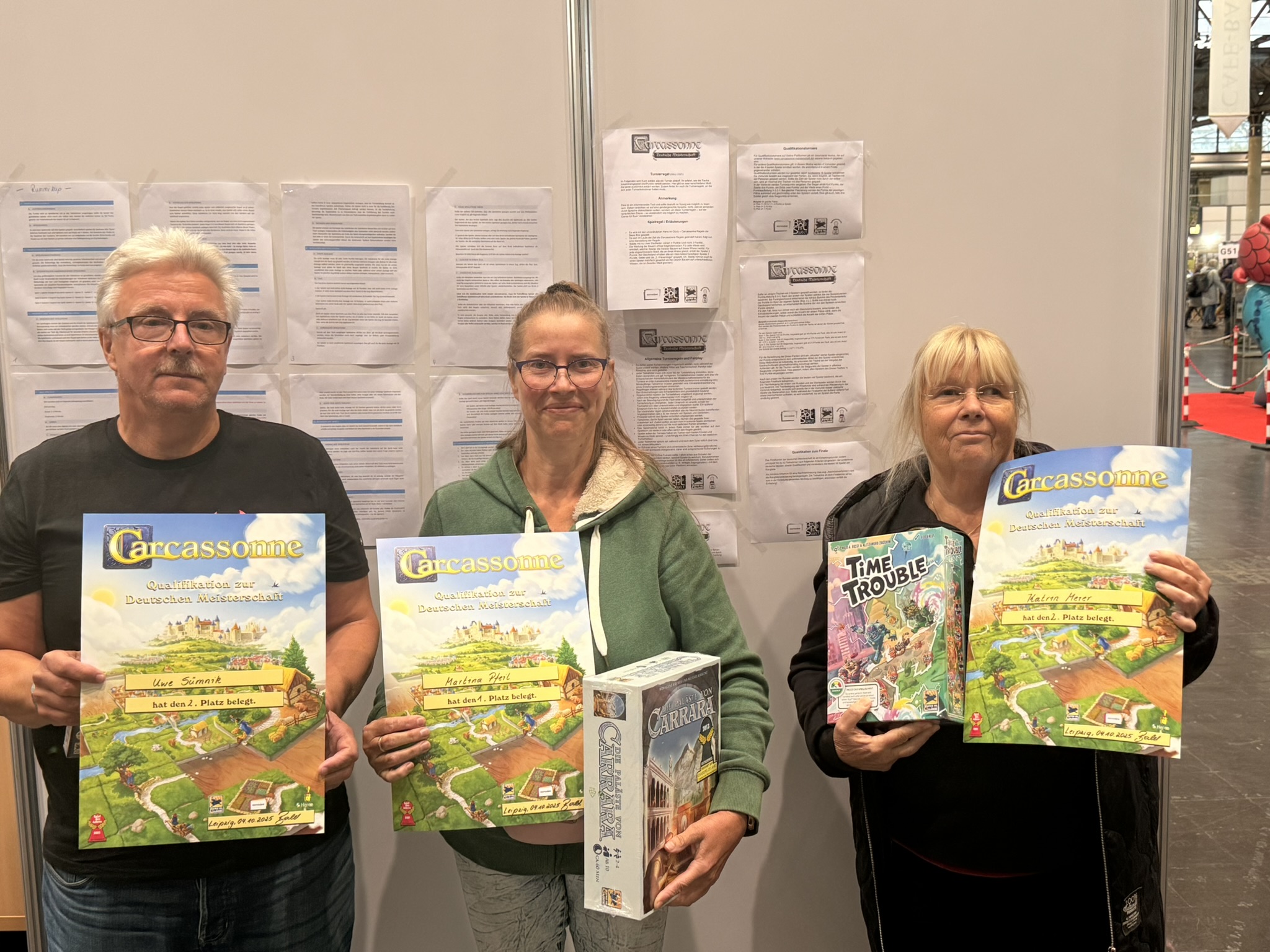 Drei Personen halten Brettspiele, darunter Carcassonne und Time Trouble, auf einer Messe.