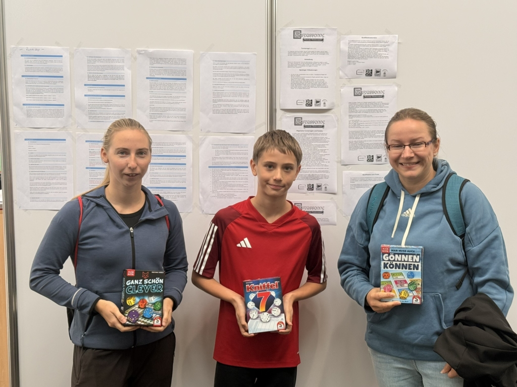 Drei Personen halten Spielverpackungen in der Hand, stehen vor einer Wand mit Informationsblättern.