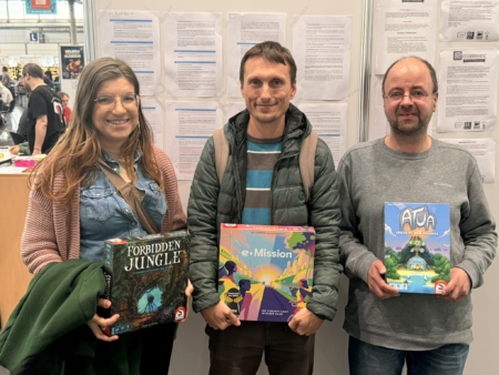 Drei Personen halten Brettspiele in der Hand, darunter 'Forbidden Jungle', 'e-Mission' und 'Atua'.