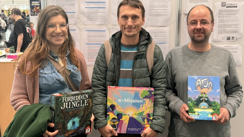 Drei Personen halten Brettspiele in der Hand, darunter 'Forbidden Jungle', 'e-Mission' und 'Atua'.