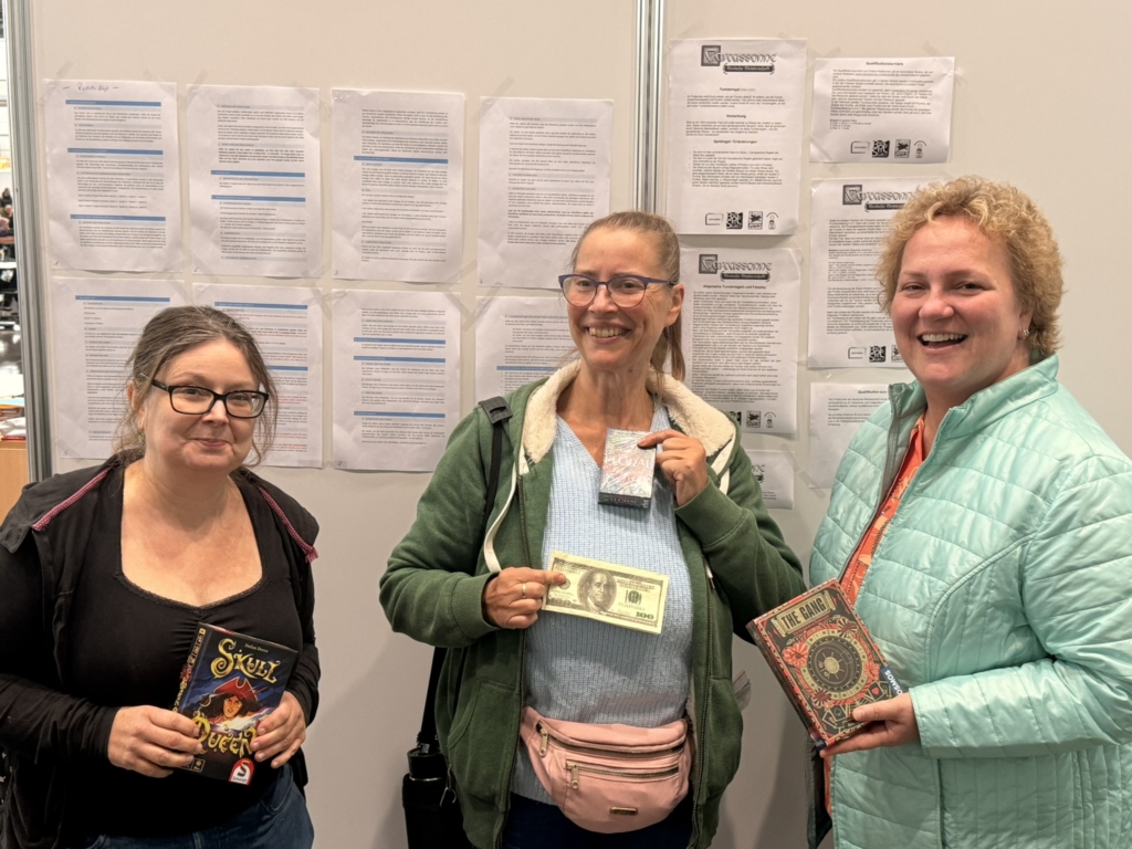 Drei Frauen halten Bücher und einen Geldschein in einer Indoor-Umgebung.