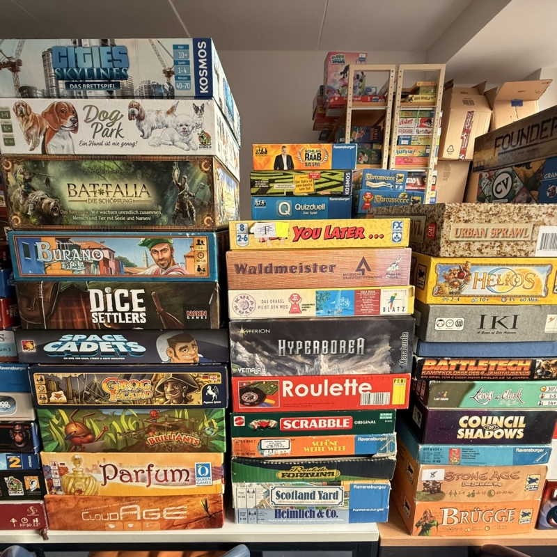 Verschiedene Brettspiele, gestapelt in einem Raum, darunter Dog Park, Scrabble und Dice Settlers.