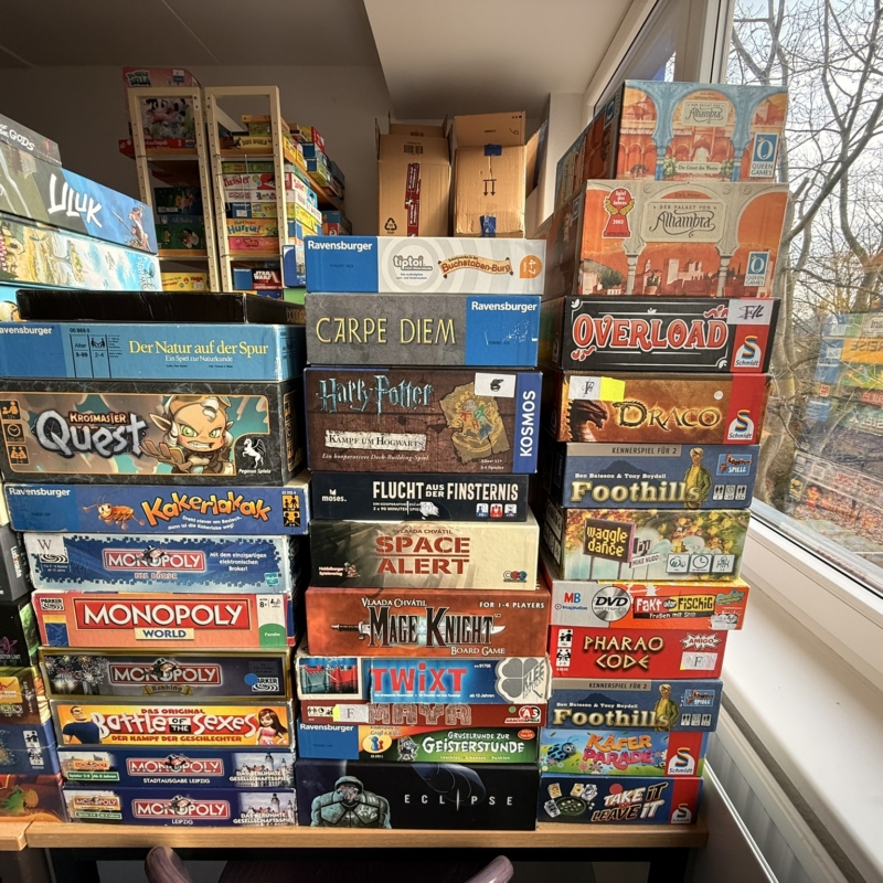 Bunte Spieleboxen gestapelt in einem Raum, darunter Klassiker wie Monopoly und Harry Potter.