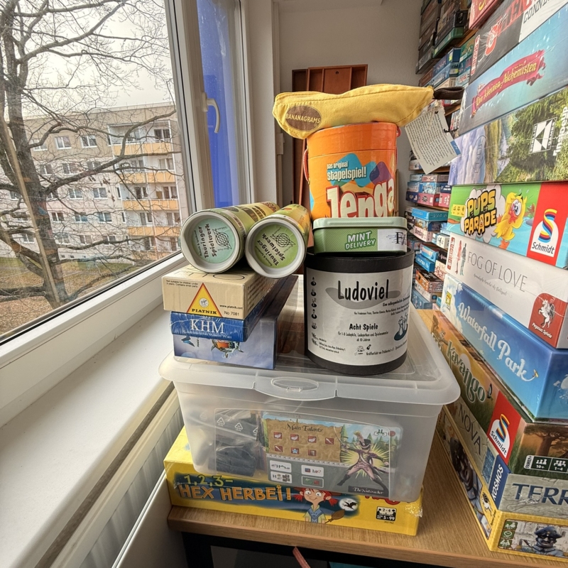 Ein Stapel von Brettspielen und Spielmaterialien auf einem Tisch neben einem Fenster.
