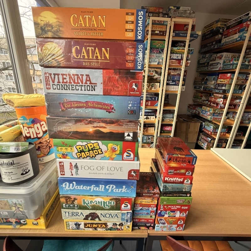 Eine Sammlung von Brettspielen, darunter Catan, Vienna Connection und Waterfall Park, auf einem Tisch gestapelt.
