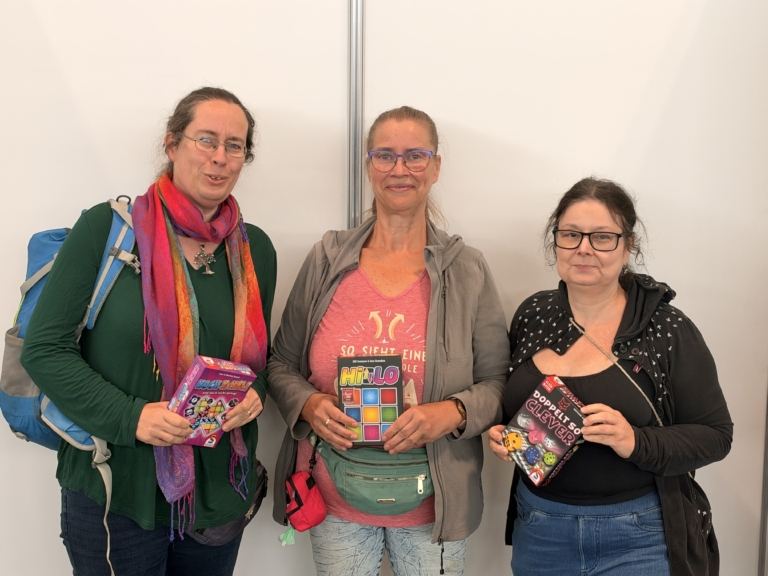 Drei Frauen präsentieren Brett- und Würfelspiele: „Noch X-mal!“ links, „Hitster“ in der Mitte und „Doppelt so clever“ rechts.