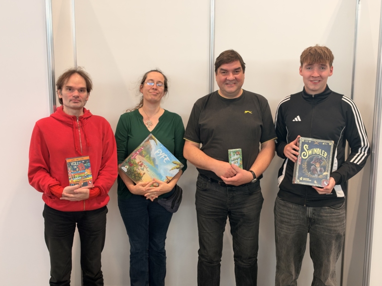 Vier Personen präsentieren Brettspiele, darunter die Titel „Voll die 18“, „EVE“ und „Swindler“, vor einer hellen Trennwand.