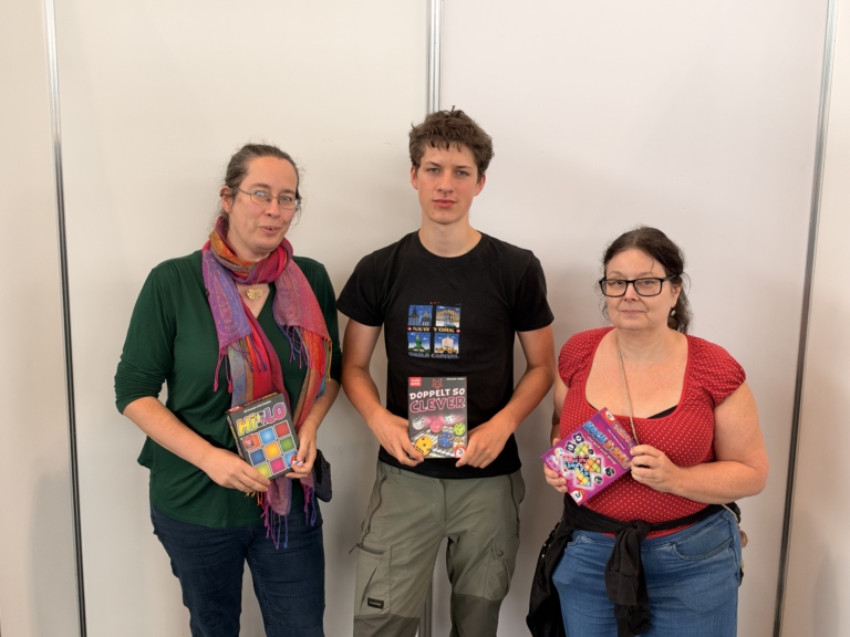 Drei Personen präsentieren Brett- und Denkspiele, darunter HILO, Doppelt so clever und ein buntes Spiel mit Zahlen und Symbolen.