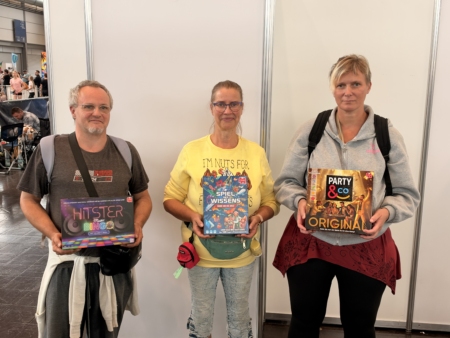 Drei Teilnehmende der Hobbymesse 2026 zeigen Gesellschaftsspiele: „Hitster Music Bingo“, „Spiel des Wissens“ und „Party & Co Original“.