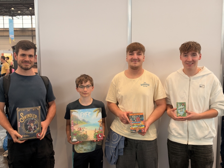 Auf einer Spielemesse zeigen vier Männer die Brettspiele Swindler, EYE, Voll die 18 und Leaf It! vor einer hellen Standwand.