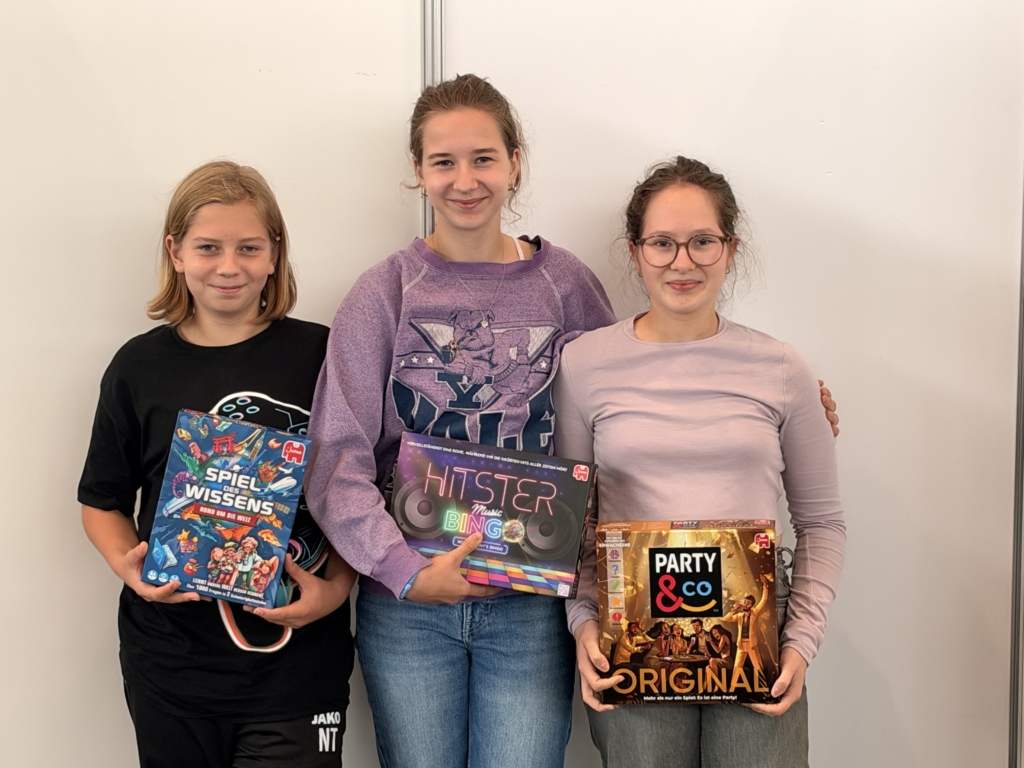 Drei junge Frauen halten Spieleboxen mit „Spiel des Wissens“, „Hitster Music Bingo“ und „Party & Co Original“ bei der Hobbymesse 2026.