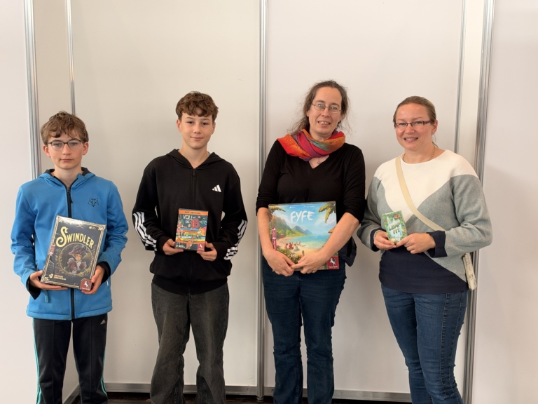 Vier Personen präsentieren die Brettspiele Swindler, Volle die 18, EYE und ein kleines Spiel vor einer hellen Wand.